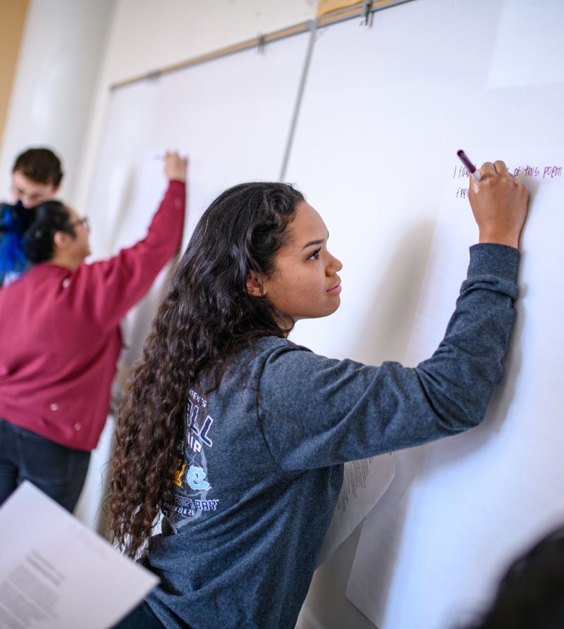 Students writing on a whiteboard during a classroom activity. A young woman in the foreground is focused as she writes with a marker.